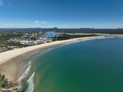 A wide drone aerial view of Laguna Bay, Noosa Spit and Main Beach with the Glass House Mountains visible on the horizon.