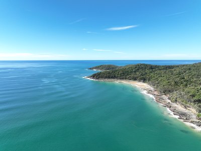 A close aerial view of a secluded Noosa National Park cove with rocky headland, dense native vegetation and turquoise ocean waters.