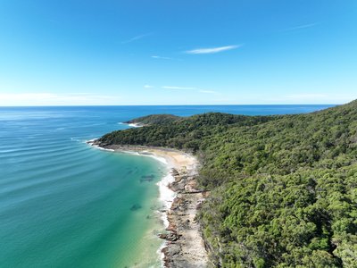 Aerial view of a hidden Noosa National Park cove with rocky sandstone cliffs, dense native vegetation and turquoise ocean waters.
