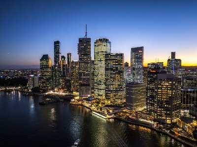 Aerial view of Brisbane CBD towers glowing at dusk with the Brisbane River in foreground under a twilight sky.