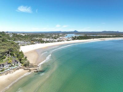 A medium altitude aerial view of Noosa Main Beach with the town visible in the distance, including buildings and vegetation.
