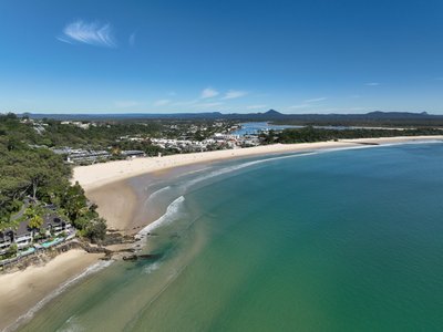 Aerial drone shot of Noosa Main Beach, showcasing the crescent shoreline and surrounding township under a clear blue sky.