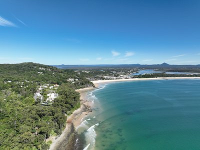 A wide aerial view of Noosa from offshore, showcasing the rocky headland and Main Beach with Sunshine Coast hinterland in the background.