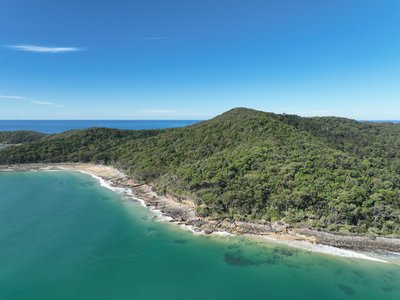 Aerial view of Noosa National Park showing dense rainforest-covered coastal hill with rocky shoreline and turquoise ocean.