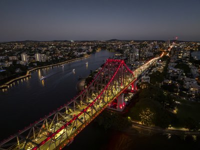 Aerial night view of the illuminated Story Bridge spanning Brisbane's river, with cityscape and suburbs visible in the background.