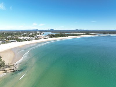 Aerial view of Noosa Main Beach arc with Noosa township, Noosa River mouth and hinterland visible under bright midday sun.