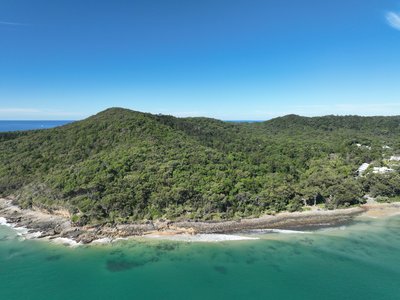Aerial view from offshore showing two forested Noosa National Park hills with rocky base, native vegetation and turquoise water.