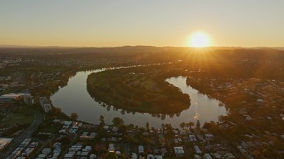 Aerial view of the Brisbane River meander at sunset with suburbs and mountain range behind.