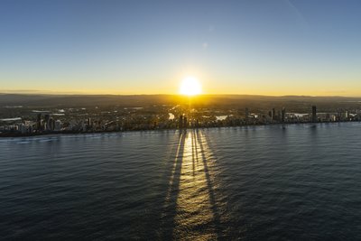 Aerial view of Gold Coast skyline with dramatic tower shadow patterns on ocean surface during sunrise.