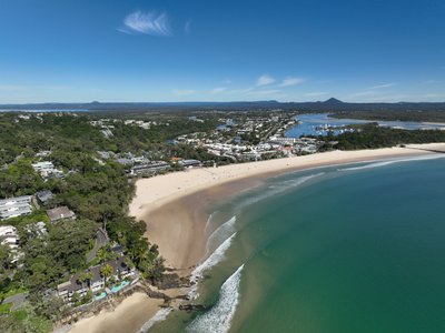 A wide aerial view along Noosa Main Beach looking south with the town, national park headland and river estuary below.