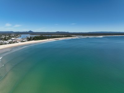 A wide aerial view of Noosa Main Beach, the mouth of the Noosa River and Noosa Spit with the Glass House Mountains visible in the background.