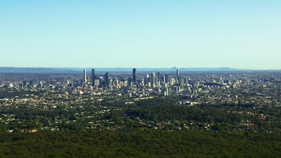 A wide aerial view of Brisbane CBD skyline with surrounding suburbs and mountains on the horizon.