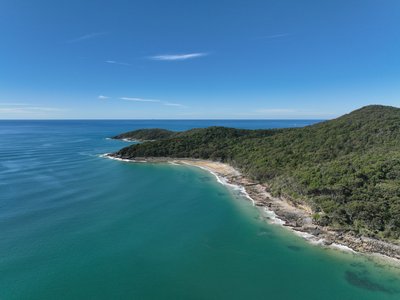 Aerial drone view of Noosa National Park's rocky headland with native rainforest-covered hills and pristine turquoise Pacific Ocean.