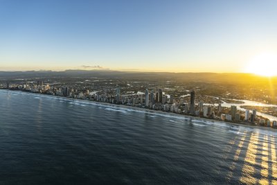 Aerial view of the Gold Coast coastline at sunrise, with high-rise buildings and hinterland mountains in the background.