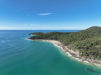 Aerial view of Noosa National Park headland showing two forested peaks, rocky coastline and a small secluded sandy beach.