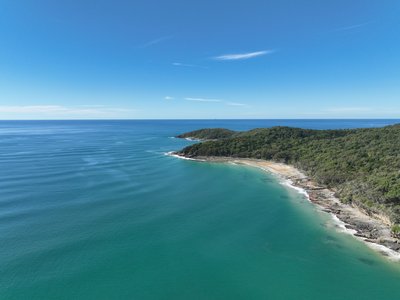 Aerial view of Noosa National Park rocky headland with a secluded cove, dense rainforest and clear turquoise waters of the Pacific Ocean.