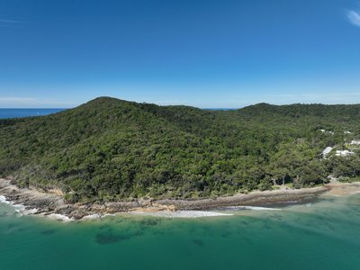 A drone view of Noosa National Park's coastal hills with dense native forest, rocky shoreline and calm turquoise ocean.