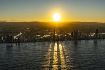 Aerial photograph of the Gold Coast sunrise with Q1 Tower silhouetted against a glowing sky, casting long shadows across the ocean.