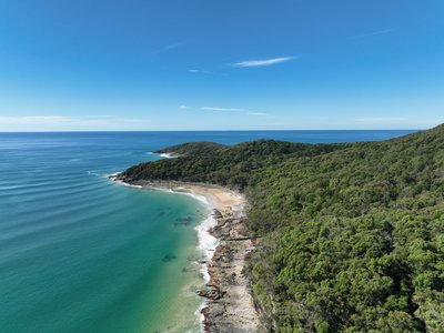 Aerial view of a remote Noosa National Park cove with rocky coastal cliffs, dense rainforest and a small secluded sandy beach.